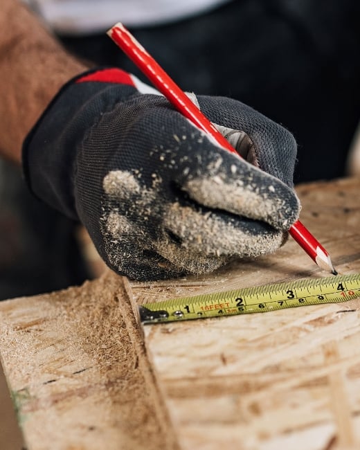 Man in black glove covered in wood shavings marking a measurement on a wooden surface